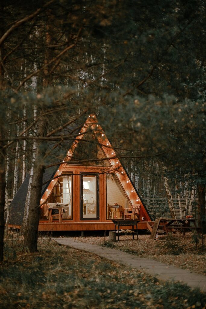 Маленький A-frame дом 36 м² с подвесным креслом, шесть одинаковых, до 4 гостей: дом вечером в лесу