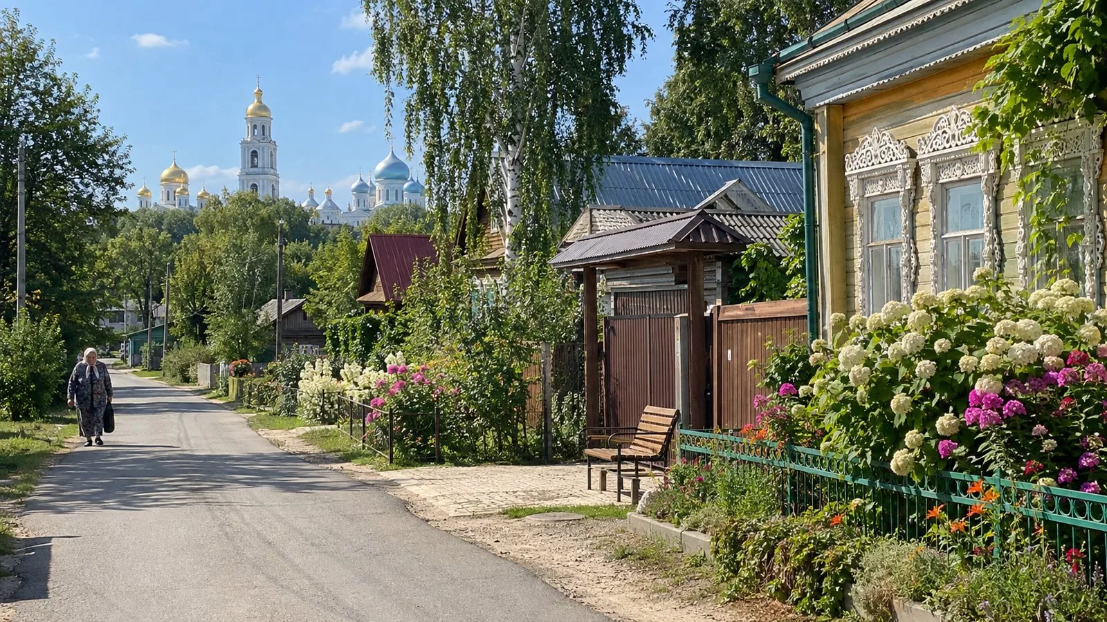 Улица Дивеева летом: деревянные дома с резными наличниками и цветниками, скамейка у калитки гостевого дома, над деревьями купола монастыря