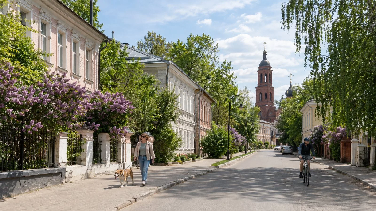 Тихая улица Клина весной: старые двухэтажные дома, цветёт сирень, впереди кирпичная колокольня, женщина с собакой и велосипедист