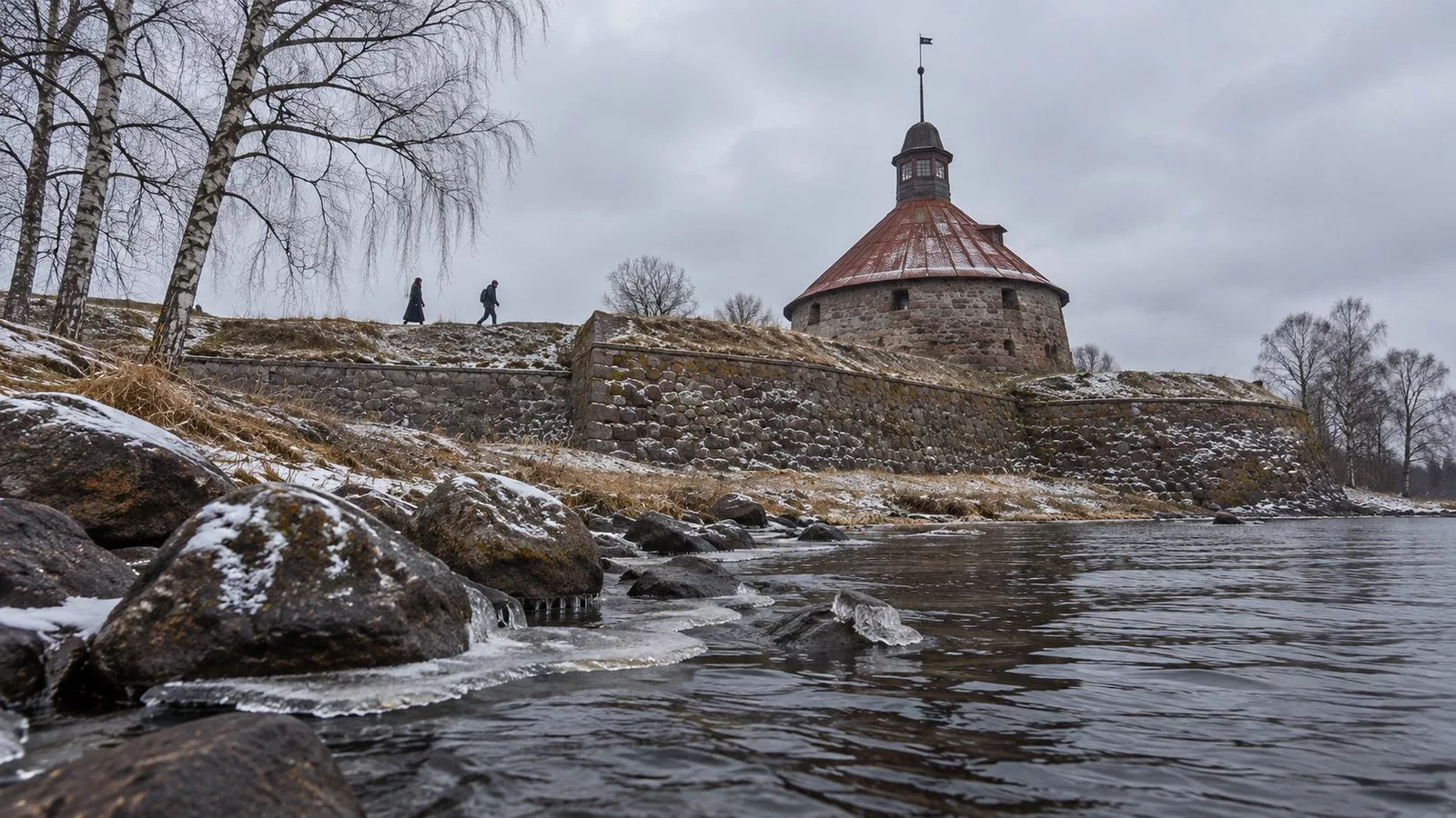 Круглая каменная башня крепости Корела над Вуоксой поздней осенью, первый снег на камнях, по валу идут двое