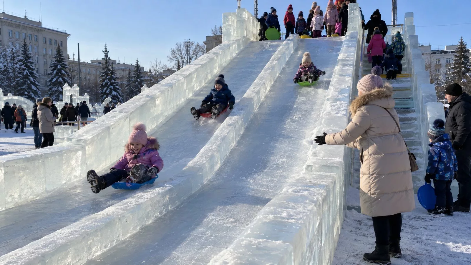 Дети съезжают с высокой ледяной горки на пластиковых ледянках на городской площади днём, внизу ждёт мама, на ступеньках очередь детей, морозный солнечный день
