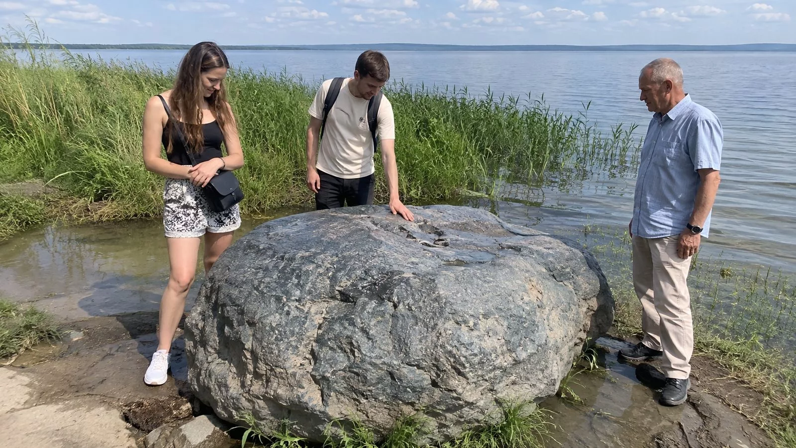 Молодая пара и пожилой мужчина стоят у большого серо-голубого валуна на берегу озера среди камыша, один касается камня