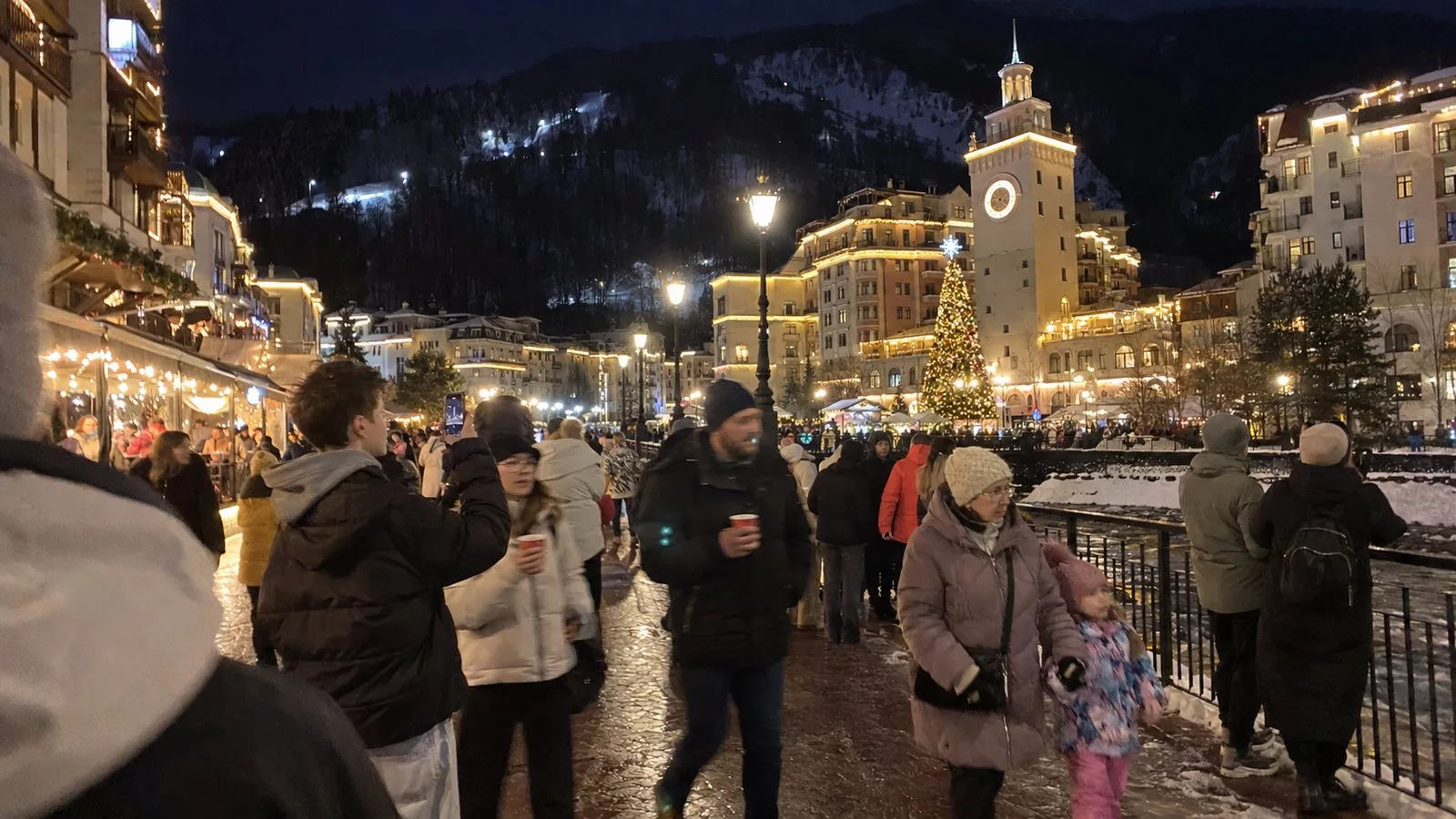 Вечер на набережной Мзымты на Розе Хутор: толпа отдыхающих в зимних куртках, наряженная ёлка у часовой башни, гирлянды на домах, подсвеченные склоны над курортом