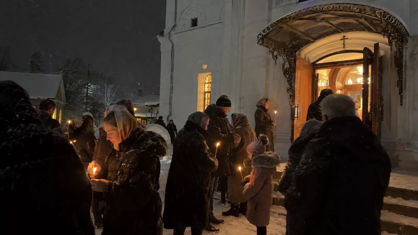 Люди со свечами стоят в снегу у белого каменного храма в рождественскую ночь, из открытых дверей храма падает тёплый свет, идёт снег