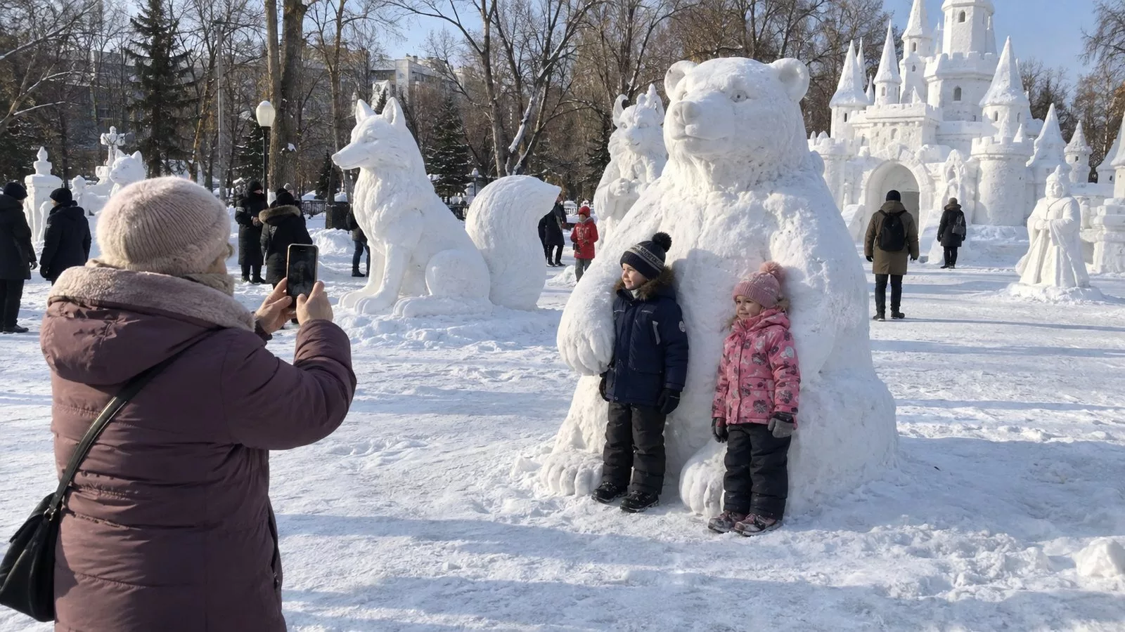 Большие белые снежные скульптуры сказочных героев и зверей в городском парке днём, бабушка фотографирует внуков рядом со снежным медведем, утоптанный снег