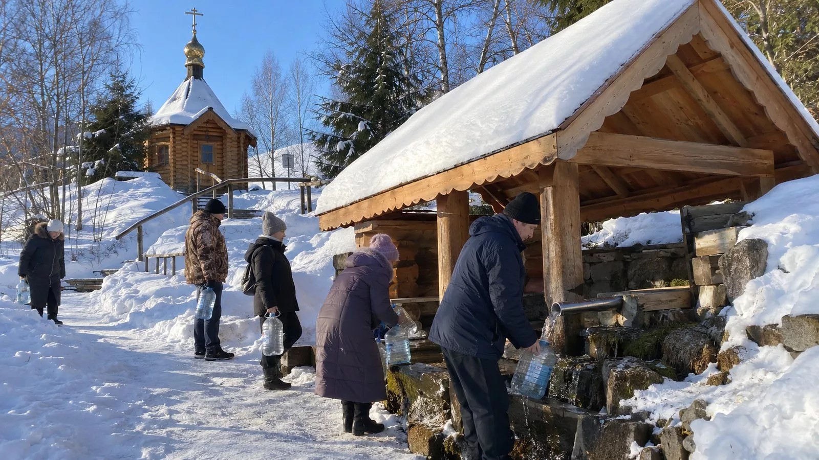 Люди разного возраста набирают воду в пластиковые бутылки из трубы у святого источника с деревянной часовней и крытой купальней, вокруг сугробы, солнечный морозный день